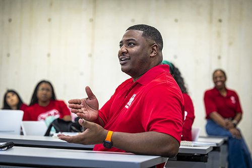 Black man in class sitting with his hands open wearing a red shirt