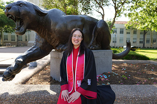 Student in regalia sitting in front of a cougar statue