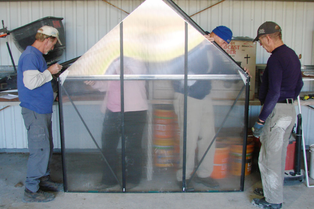Nearly assembled greenhouse with transparent panels, as multiple individuals work together to secure it.